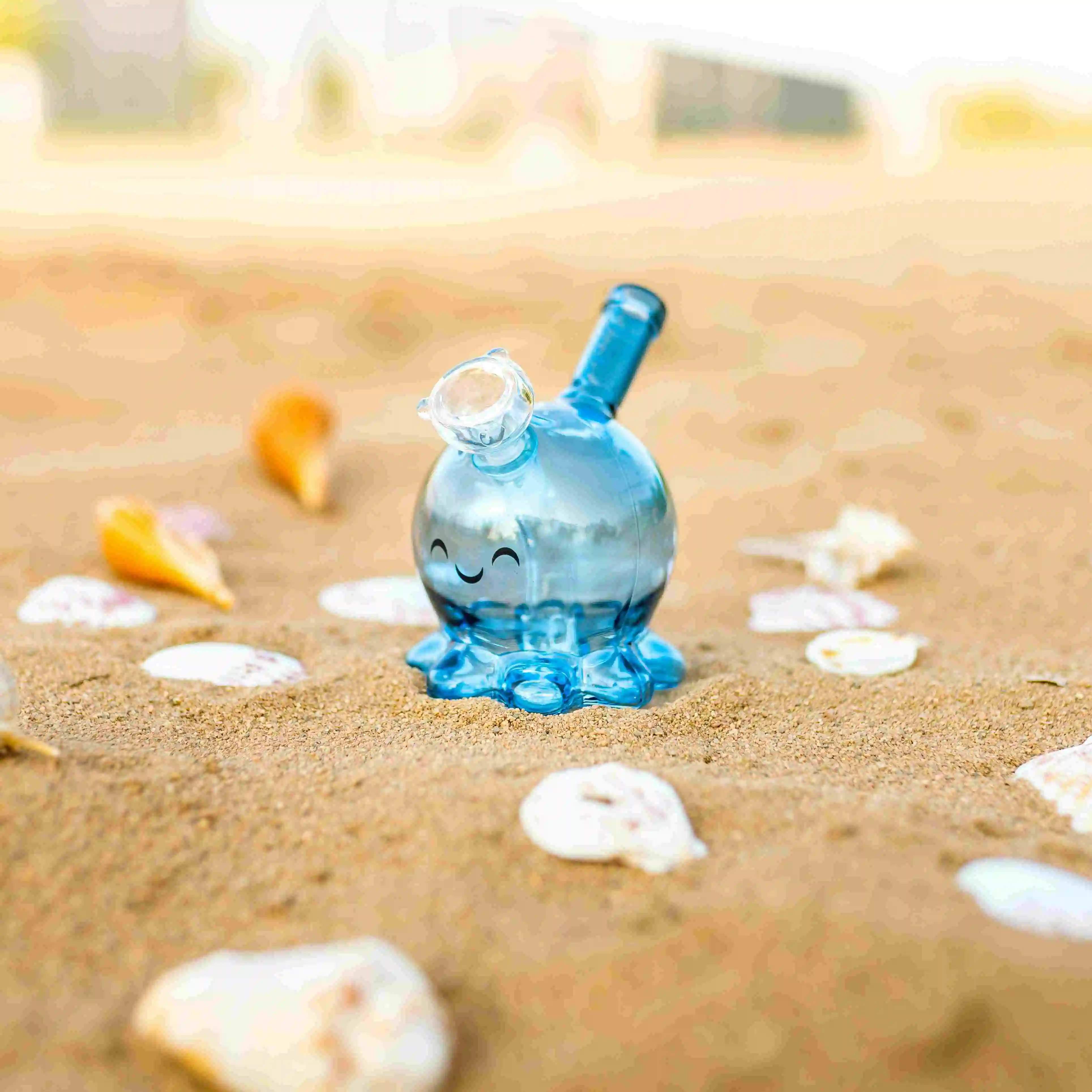 Small blue glass bong with a smiley face on the front placed on a beach with shells and sand