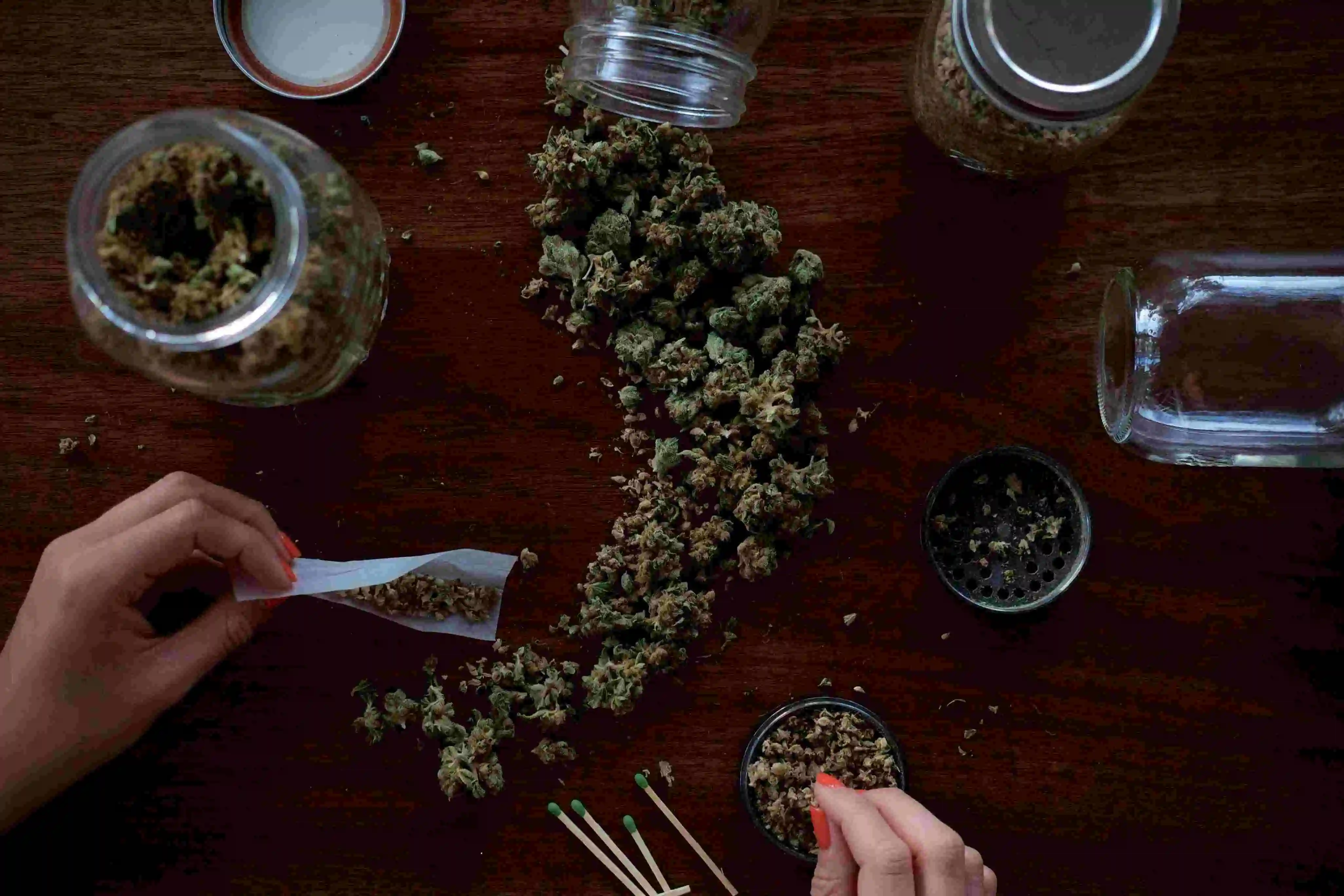 an assortment of jars with cannabis buds falling out and a pair of hands using the weed to roll a joint